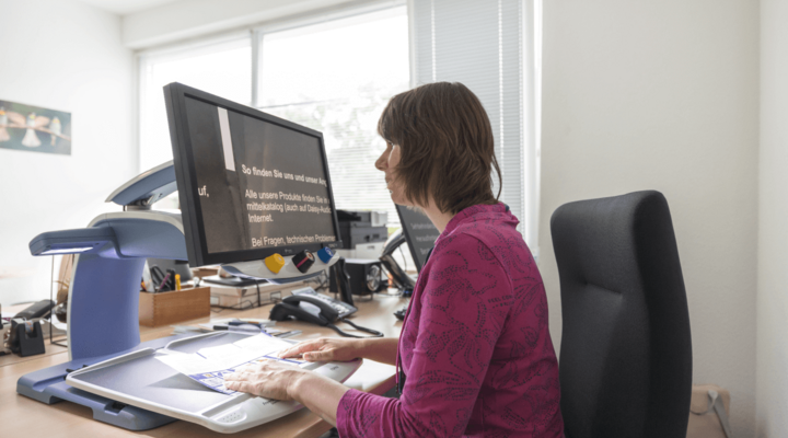 Eine Frau sitzt an einem Schreibtisch und hat ein übergroßen Bildschirm vor sich, auf dem die Schrift sehr groß abgebildet ist.