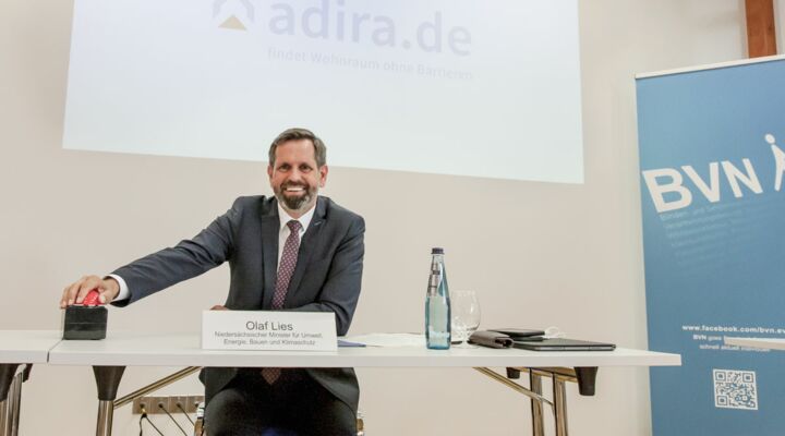 Olaf Lies drückt sitzend auf roten Buzzer. Hinter ihm an der Wand erscheint die Startseite von adira.de