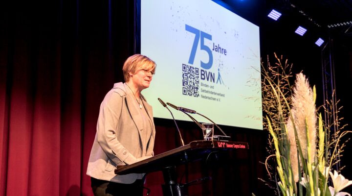 Frau Wernstedt hat halblange Haare mit blonden Strähnen, die in die Stirn fallen. Sie trägt eine Brille, eine Kette, ein beigefarbenes Sakko und eine dunkle Hose
