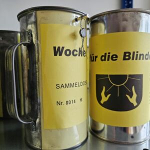 Zwei Sammeldosen sind im Vordergrund. Sie tragen ein gelbes Schild mit der Aufschrift Woche für die Blinden.