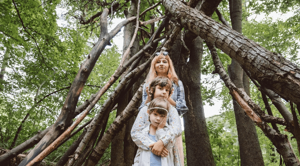 In einem Wald stehen an einem Holzunterstand eine Mutter mit zwei Kindern. Die Mutter trägt eine Brille auf dem Kopf und die Kinder haben Haarreifen.