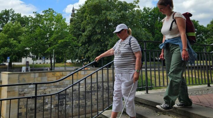 Die Klientin geht am Langstock eine Treppe herunter, die Reha-Lehrein geht nah hinter ihr und schaut auf die Stockführung. Die Klientin trägt ein weißes Cappi, sie hält sich am Geländer fest. Im Hintergrund ist ein Flussufer und ein Stadtprofil.
