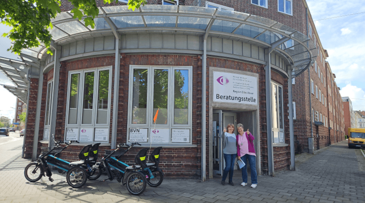 Zwei Frauen stehen vor der Beratungsstelle Elbe-Weser. Sie halten die Daumen nach oben. Vor der Hauswand steht ein Tandemrad.