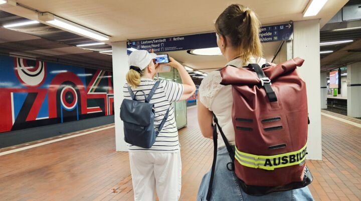 Hinteransicht von zwei Personen. Die Klientin steht links. Sie hält ihr Handy auf ein U-Bahn-Schild, auf dem die Fahrtrichtung beschrieben ist. Die Reha-Lehrerin steht vorne rechts. Auf ihrem Rucksack steht die Aufschrift Ausbilderin.