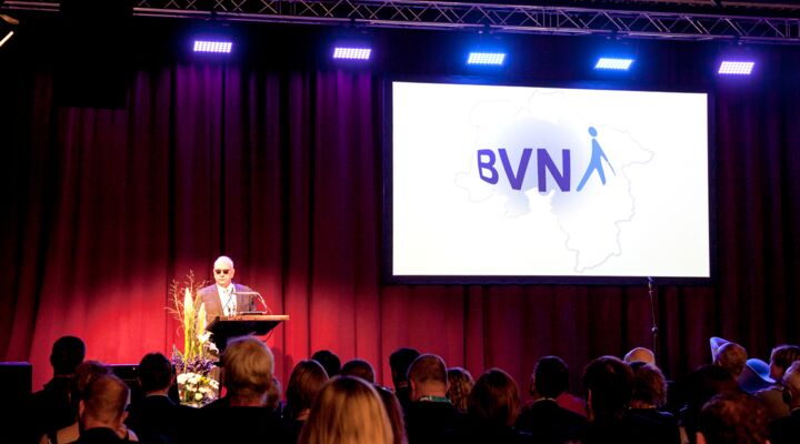 Hans-Werner Lange am Rednerpult, auf dem drei Mikrofonen stehen. Davor ein großes Blumengesteck, hinter ihm der große rote Vorhang und die Projektion des BVN-Logos in den Grenzen Niedersachsens