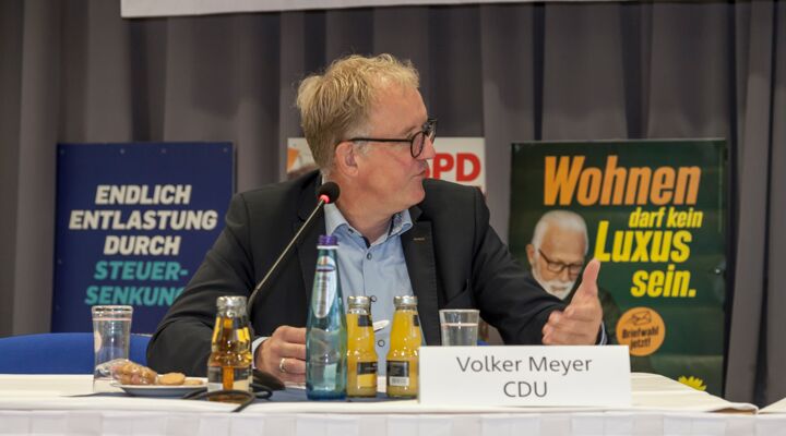 Volker Meyer sitzt am Podiumstisch. Während er spricht, schaut er nach links und unterstreicht das Gesagte mit einer Geste seiner linken Hand. Er hat blondes, Haar, trägt eine Brille, ein hellblaues Hemd, und darüber ein dunkles Sakko.