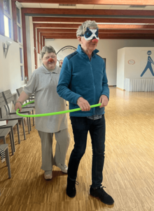 Eine Frau und ein Mann mit Clownsnasen stehen in einem Hula-Reifen und gehen durch einen großen Raum.