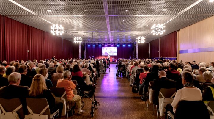 Blick aus dem hinteren Saalbereich in Richtung Bühne, auf der Corinna May steht und singt. Die Gäste in den voll besetzten Reihen sind von hinten zu sehen