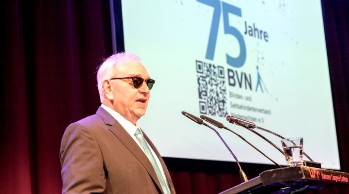 Herr Lange steht am Rednerpult. Silbriges Haar, er trägt eine Sonnenbrille, ein braunes Sakko und die hellblaue BVN-Krawatte