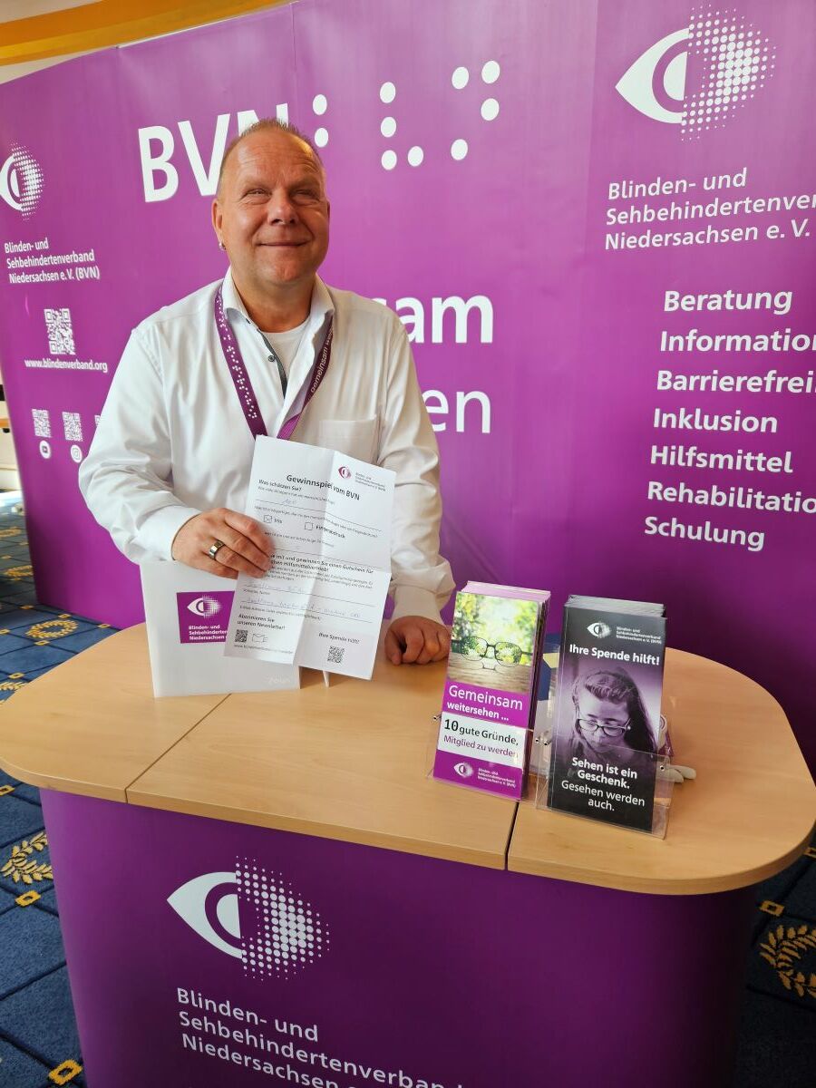 Ein Mann im weißen Hemd hält einen Zettel in die Kamera. Er steht an einer purpurfarbenen Messewand und lehnt auf einen Tresen, an dem Flyer stehen.
