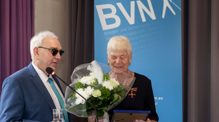 Herr Lange spricht ins Mikrofon. Frau Neumann hat eine blonde Kurzhaarfrisur, Ohrringe, sie trägt ein langes, schwarzes Kleid mit dem Bundesverdienstkreuz. In einer Hand hält sie einen Blumenstrauß, in der anderen die Ehrenmitgliedschaftsurkunde.