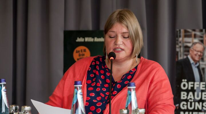 Katrin Mangelsdorf-Lüdeke sitzt am Vorstandstisch. In ihrer rechten Hand hält sie ein Blatt. Sie spricht ins Mikrofon. Sie trägt eine schwarze Bluse mit leuchtend roten Punkten, einen schwarzen Rock und eine halblange hellrote Strickjacke