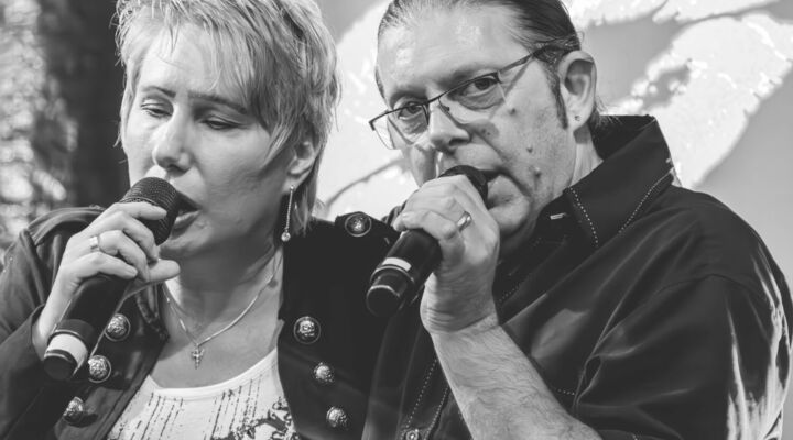 Eine Frau und ein Mann sind im Porträt als Schwarz-Weiß-Aufnahme zu sehen. Sie haben beide ein Mikrofon in der Hand und singen. Die Frau ist blind, es ist Corinna May.