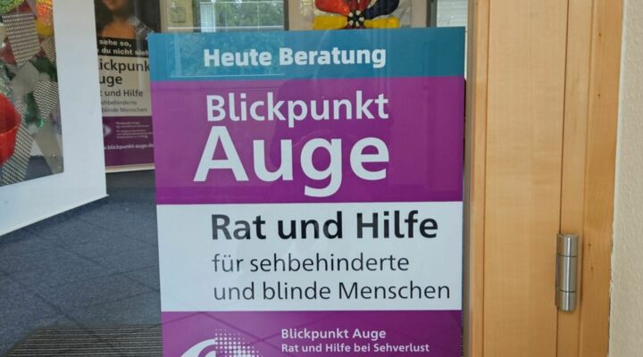 An einer Holz-Glas-Tür hängt ein Schild mit Blickpunkt-Auge-Aufschrift. Im Hintergrund ist im Raum ein Roll-up zu erkennen.