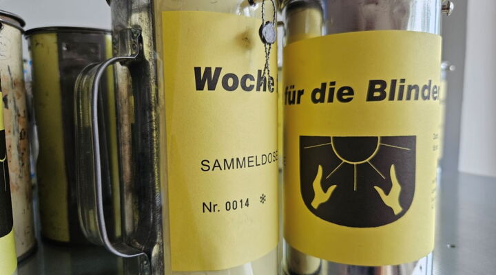 Zwei Sammeldosen sind im Vordergrund. Sie tragen ein gelbes Schild mit der Aufschrift Woche für die Blinden.