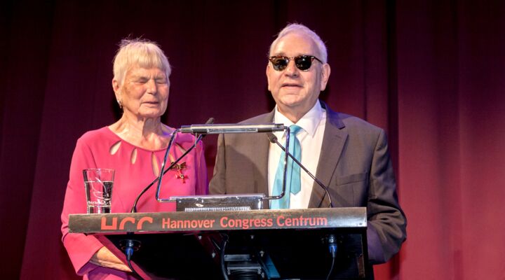 : Frau Neumann in einem roten Kleid, sie trägt das Bundesverdienstkreuz, das ihr vor Jahren verliehen wurde. Hans-Werner Lange rechts neben ihr, graues Sakko, weißes Hemd, blaue BVN-Krawatte. Er trägt eine Sonnenbrille und spricht gerade