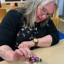 Eine Frau mit grauen Haaren und einer Brille hält eine kleine Flasche über ein mit weißen Kristallen gefülltem Glas.