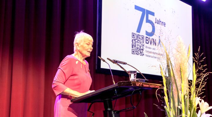 Frau Neumann, kurz geschnittene blonde Haare, rotes Kleid, steht am Rednerpult, davor ein großes Blumengesteck. Hinter ihr ein karminroter schwerer Vorhang, und die gr0ße Projektion „75 Jahre BVN“