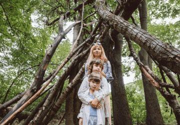 In einem Wald stehen an einem Holzunterstand eine Mutter mit zwei Kindern. Die Mutter trägt eine Brille auf dem Kopf und die Kinder haben Haarreifen.