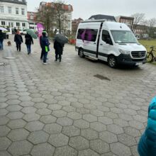 Auf einem großen Platz steht das BPA-Mobil. Es ist ein weißer Sprinter mit dem purpurfarbenen Logo. Davor stehen einige Menschen in Regenkleidung. Im Hintergrund ist eine Häuserwand zu sehen.