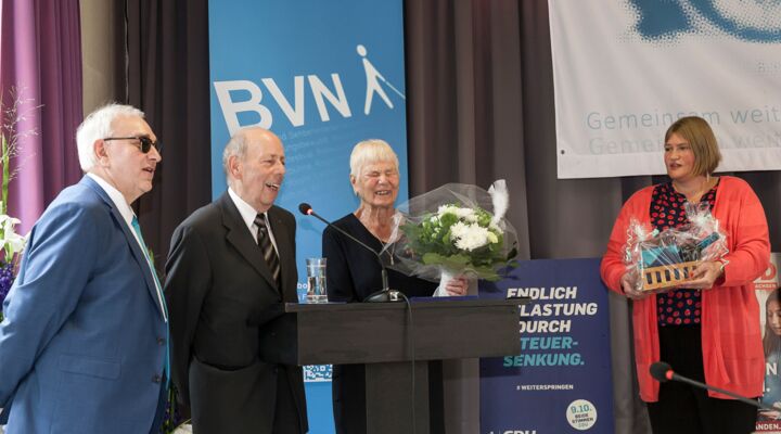 Links im Bild Hans-Werner Lange. Rechts daneben am Rednerpult spricht Manfred Barnstorf. Neben ihm Helga Neumann mit dem Blumenstrauß. Ganz rechts Katrin Mangelsdorf Lüdeke, halblange dunkelblonde Haare, schwarze Bluse mit leuchtend roten Punkten.