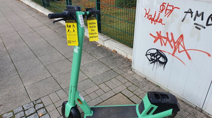 Ein Roller steht quer auf dem Gehweg. An seinem Lenker ist eine längliche gelbe Karte mit dem Text „Gelbe Karte für Falschparker“ angebracht.