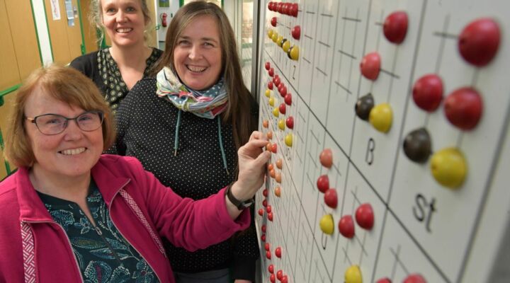 Drei Frauen lachen fröhlich in die Kamera. Sie stehen vor einer Braille-Wand. Sie tragen bunte Kleidung und sind sehr zugewandt.