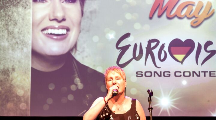 Corinna May auf der Bühne, sie trägt jetzt ein ärmelloses bräunliches Kleid mit weißen Noten, Notenschlüsseln und Sternen. Groß im Hintergrund eine Projektion zum Eurovision Song Contest und einem damals aktuellen Foto von ihr