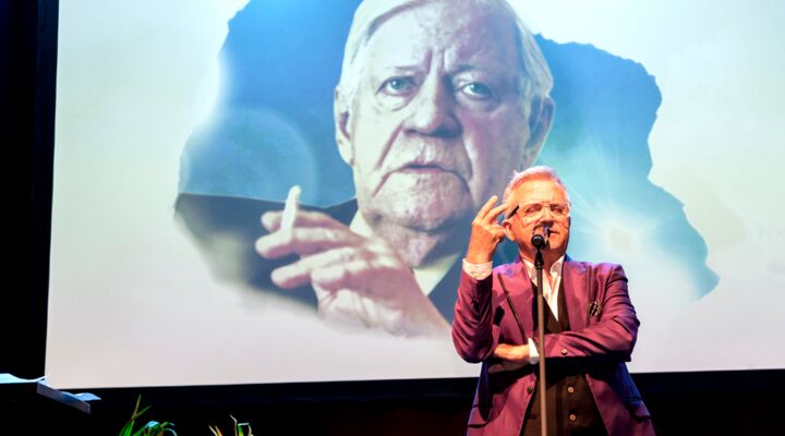 Jörg Knör steht auf der Bühne, linken Arm vorm Körper verschränkt, den rechten erhoben. Zeige- und Mittelfinger imitieren das Halten einer Zigarette. Hinter ihm eine Projektion mit dem Konterfei von Helmut Schmidt, der eine Zigarette raucht