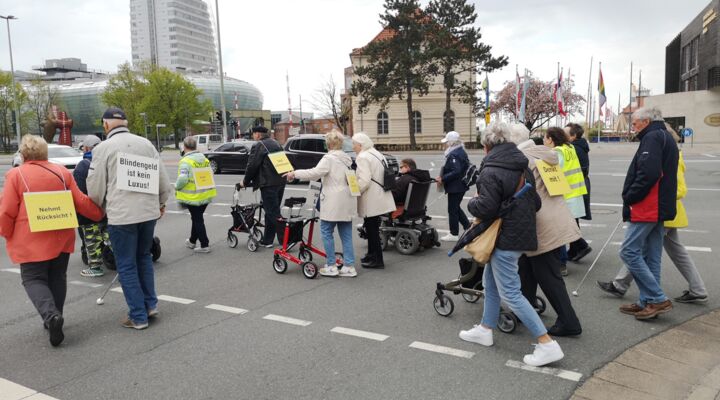 Eine Gruppe an Menschen mit und ohne Sehbeeinträchtigung geht über eine belebte Straße. Einige aus der Gruppe nutzen Gehhilfen, den Rollator oder auch den Langstock. Die Menschen tragen Schilder mit Aufschriften wie „Nehmt Rücksicht!“