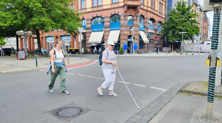 An einer Ampelkreuzung in der belebten Innenstadt überqueren Reha-Lehrerin und Klientin die Straße. Die Klientin nutzt einen Langstock und geht voran.