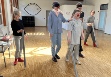 Fünf Frauen sind in einem großen Raum. Zwei stehen etwas weiter weg, drei Personen sind nah beieinander. Sie tragen Clownsnasen und zwei von ihnen Schlafbrillen. Drei Frauen haben eine Pool-Nudel in der Hand.