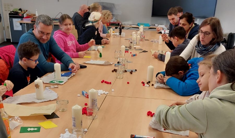 An einem langen Holztisch sitzen einige Kinder mit ihren Eltern und basteln mit weißen Kerzen, Klebe-Bällen und Wachsmustern.
