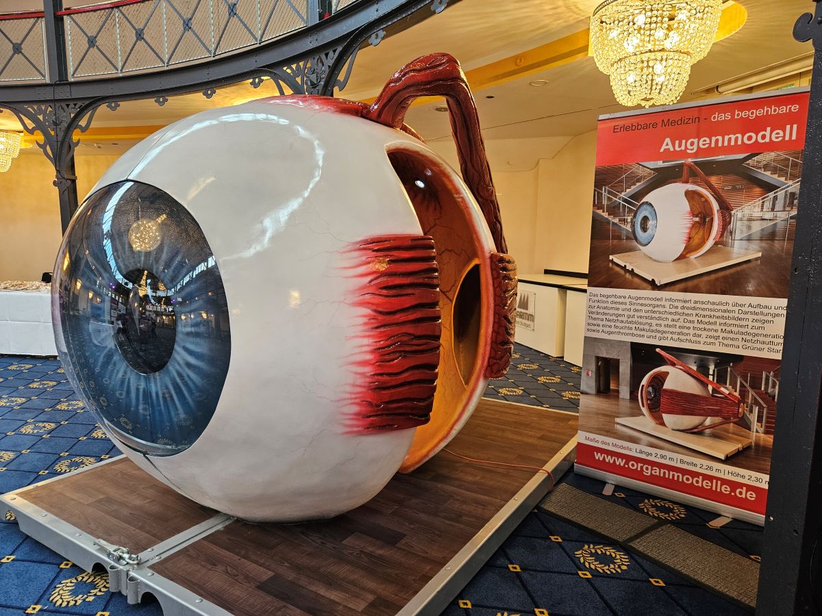 Ein großes Auge steht auf einer Holzpalette in einem großen Raum. Daneben steht ein Roll-up mit weiteren Informationen. Das Auge hat eine blaue Augenfarbe.