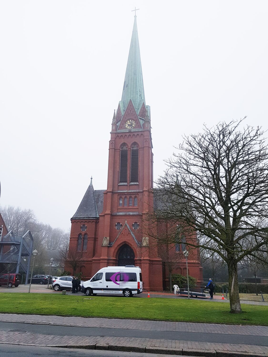 Im Hintergrund ist die große, braune Banter Kirche. Der Himmel lässt regnerisches Wetter vermuten. Auf dem Platz vor der Kirche steht der weiße Mercedes-Sprinter mit dem Logo vom BVN und dem Angebot „Blickpunkt Auge“.